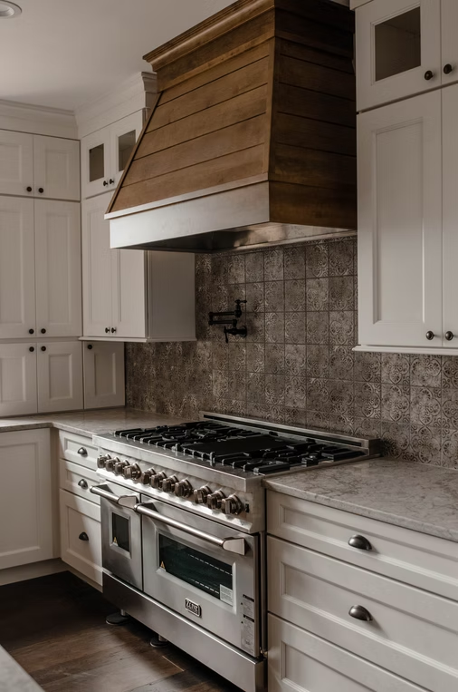 Custom kitchen with wood range hood, white cabinetry, marble counters, and patterned tile backsplash